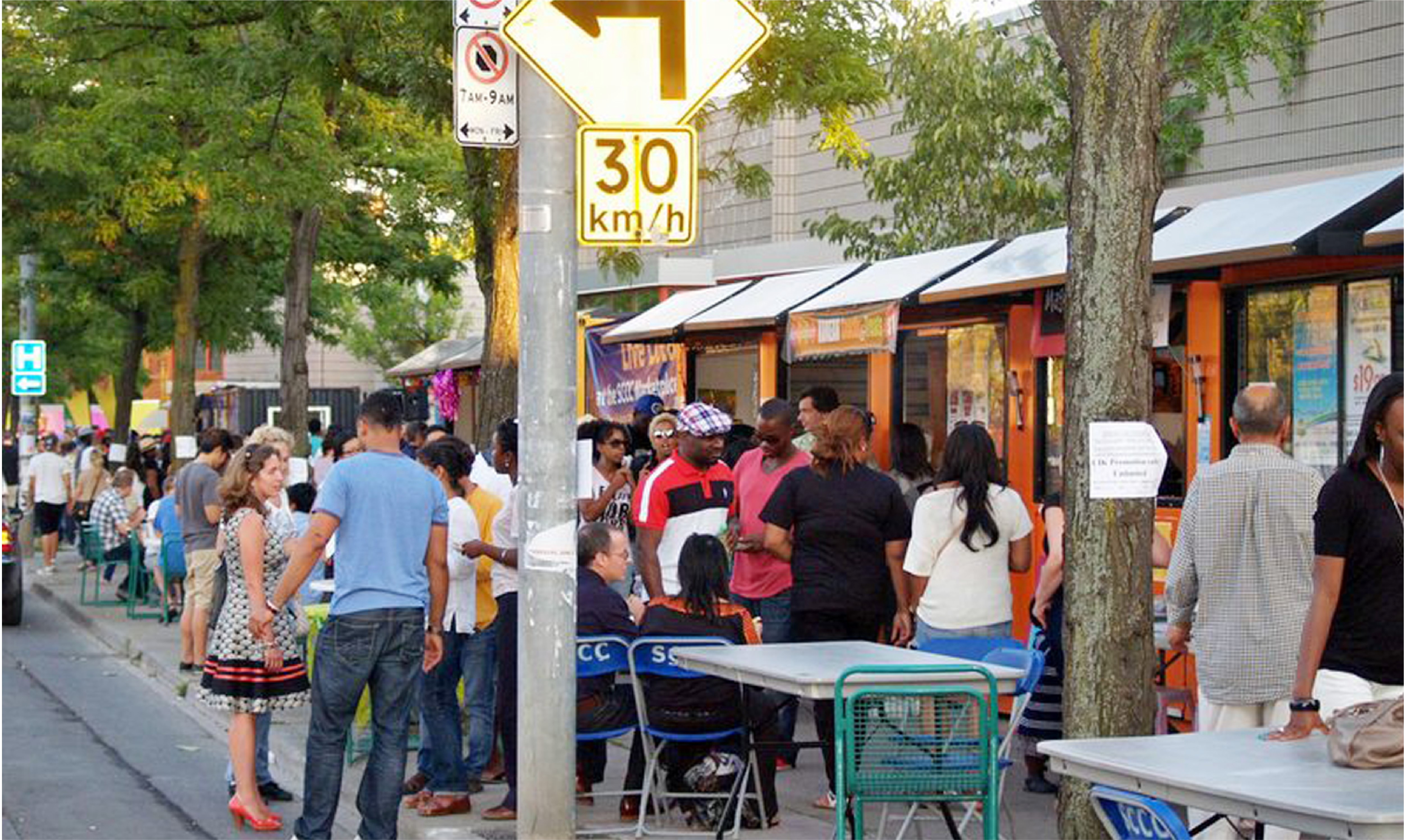 A line up of people on the sidewalk in front of several small outdoor shopping stalls.