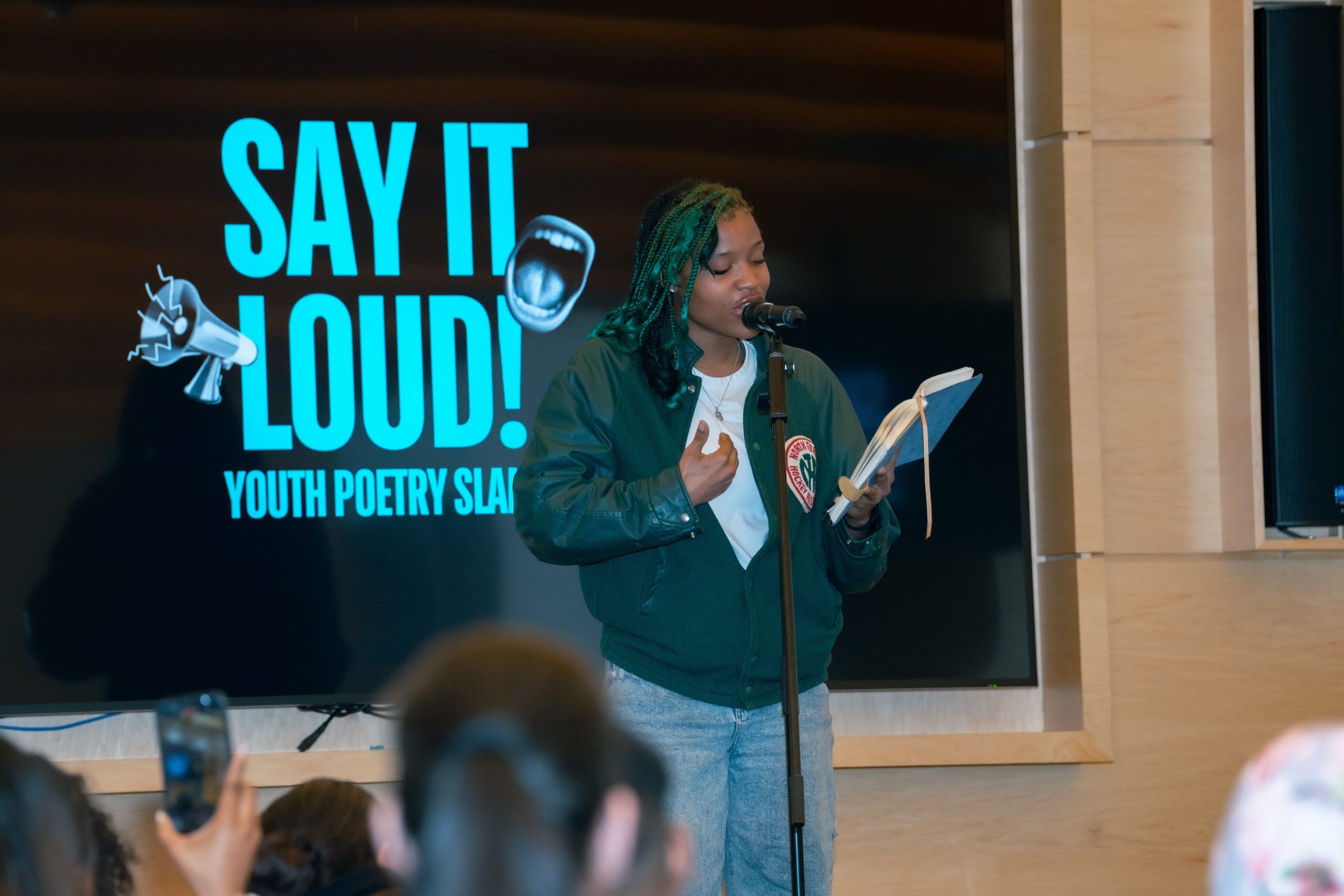 A person stands in front of a microphone, holding a book or notebook, and speaking. Behind them is a large screen displaying the text "SAY IT LOUD! YOUTH POETRY SLAM" with an image of an open mouth and a megaphone.