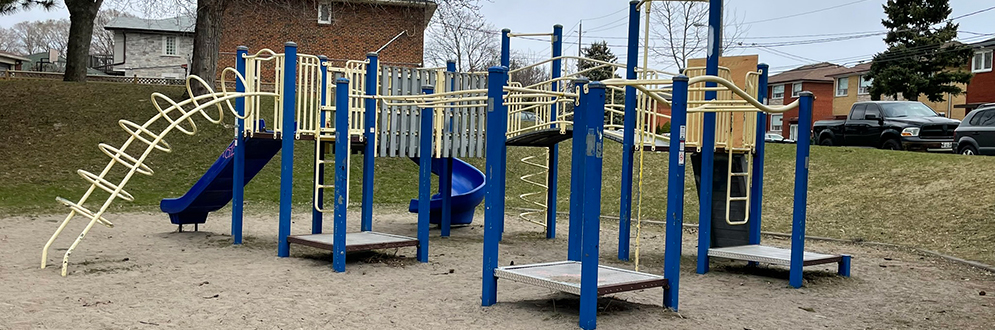 A blue and yellow playground structure at Cayuga Park sits on a sandy surface. The structure includes slides, ladders, climbing bars, and platforms. Scattered toy trucks and cars are visible in the sand. Leafless trees, residential houses, and parked cars surround the park under a cloudy sky.