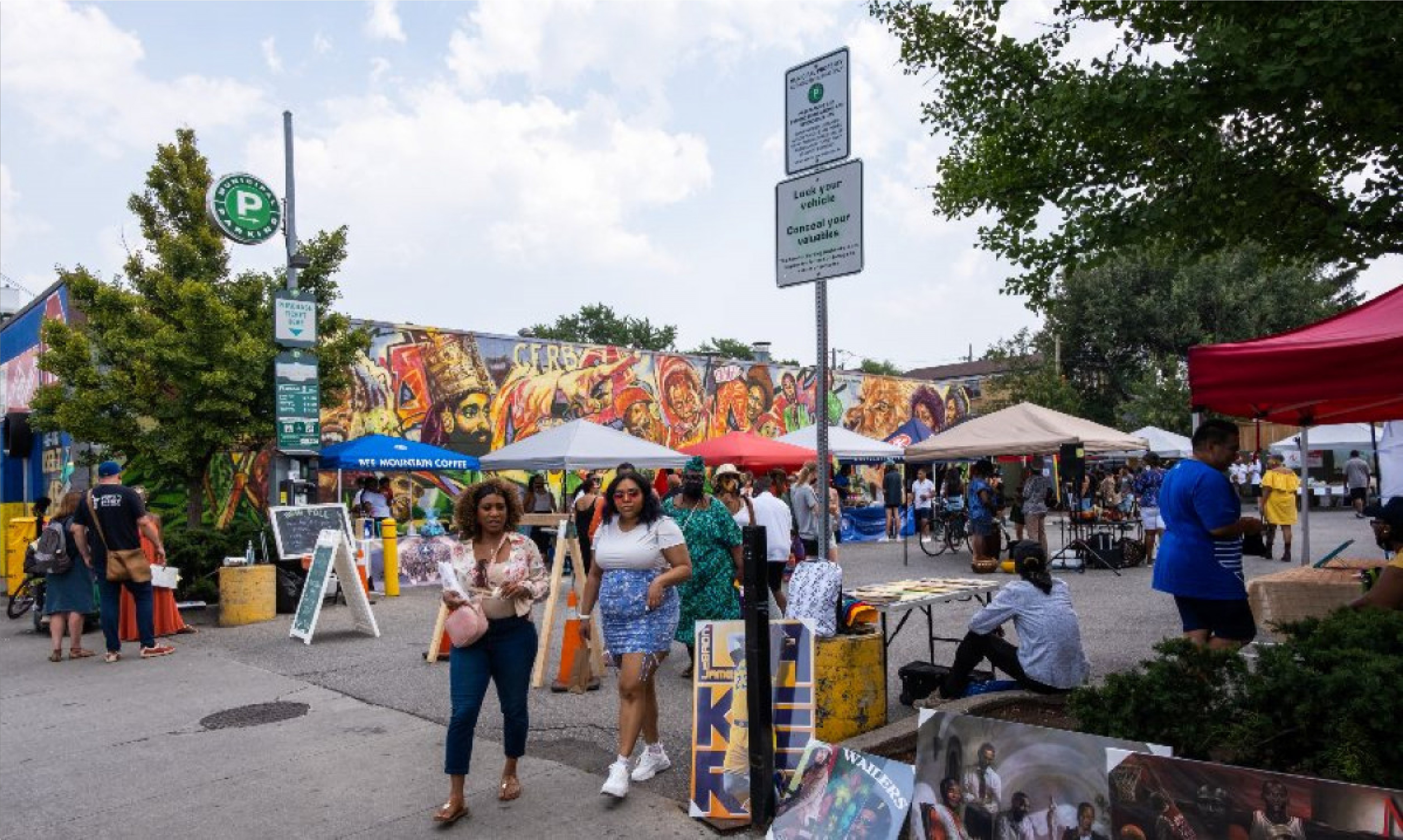 A busy market in a parking lot busy with people. A vibrant mural is painted on the building next to the parking lot.
