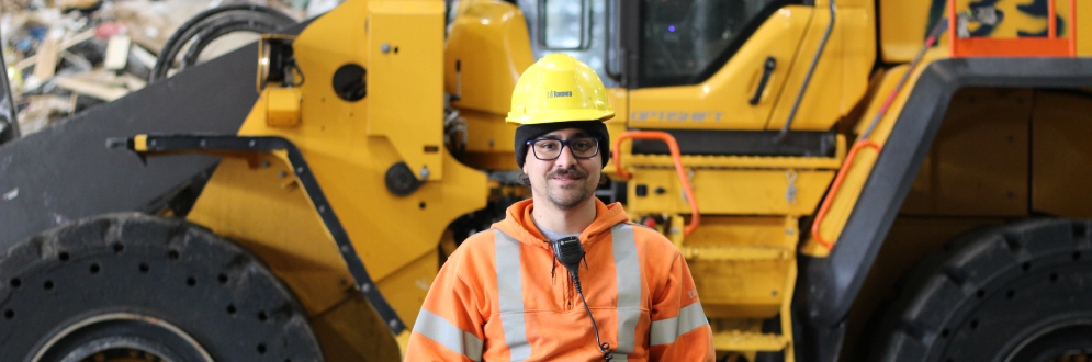 A City of Toronto worker wearing a hardhat and orange sweater, He is standing in front of a large powered vehicle.