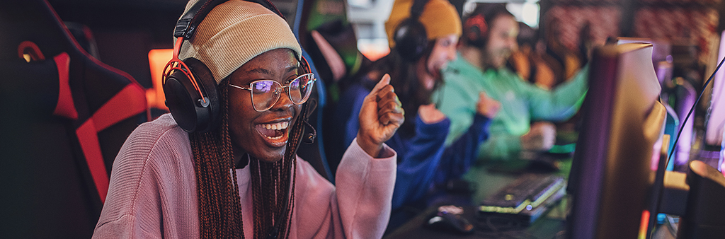 A woman sitting in a row of gamers in front of a monitor and wearing a headset with a big smile on her face.