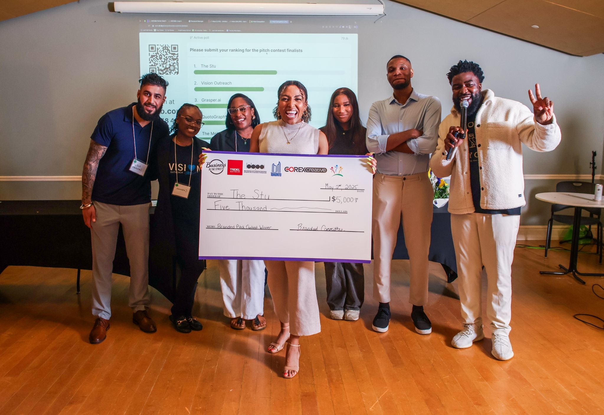A group of six people standing indoors, with one person in the center holding a large check. The check is made out to "The Sibs" for the amount of "Five Thousand" dollars.