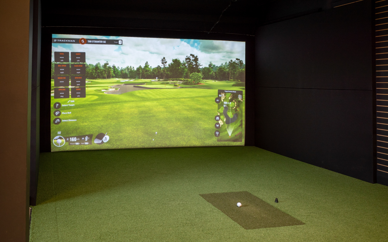 A golf simulator setup featuring a large projection screen on the wall. The floor is covered with artificial grass, and the surrounding walls are dark. A golf ball lies on the turf, ready for a player to swing.