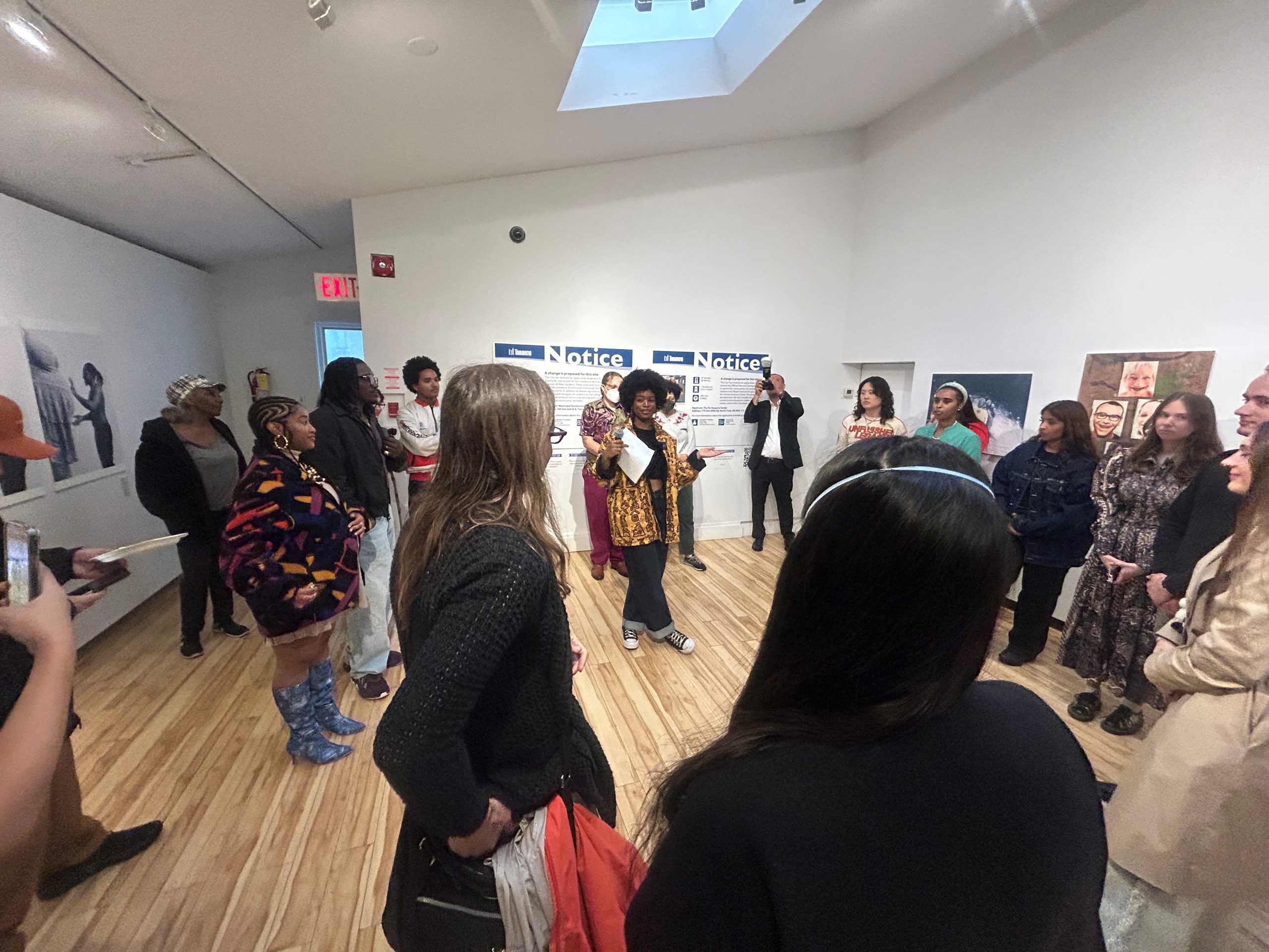 A group of people is gathered in an art gallery. The room has white walls and a wooden floor. There are several pieces of artwork displayed on the walls, including photographs and paintings. Some people are standing in a circle, engaged in conversation and listening to someone speak.