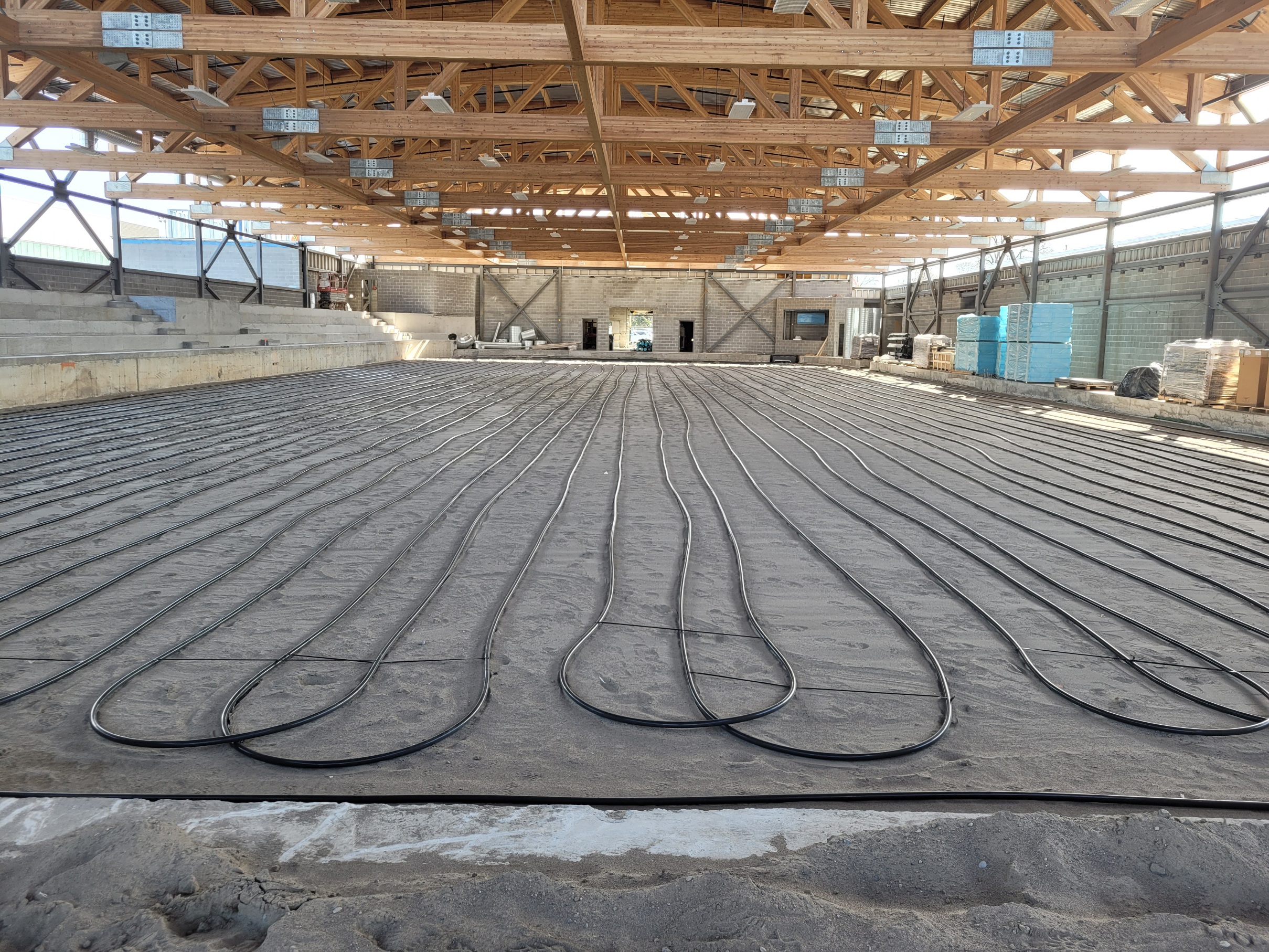 An interior view of an arena and ice rink under construction, showing the installation of refrigeration piping in progress. The large space features grey brick walls with metal scaffolding in place to support the wooden ceiling beams. At the centre where the rink will be, black tubes are laid lengthwise in parallel loops on a sandy base. To the left of the rink are concrete bleachers. Construction materials and equipment are piled along the other side of the rink.