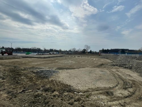 View of the Centennial Park baseball hub under construction, focusing on the drainage work being carried out on the surface to manage and collect storm water. The dirt-covered construction site features a levelled area with visible tire tracks surrounded by mounds of soil and gravel. Construction containers and equipment are scattered across the site, which is enclosed by blue fencing. In the background, there is a parking lot with vehicles and streetlights, trees and low buildings under a partly cloudy sky.