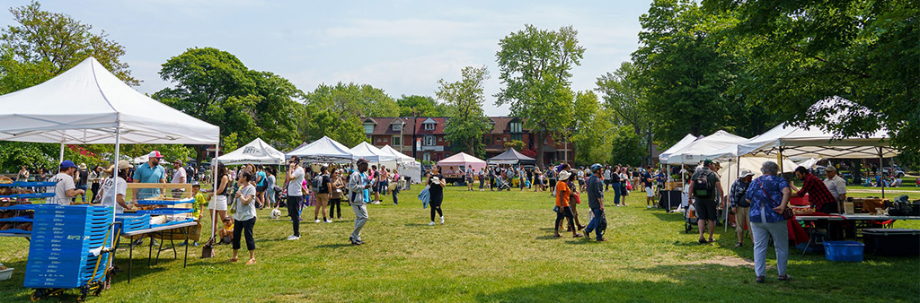 Many white tents set up in a park, people shopping at the outdoor market.