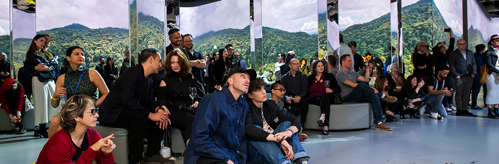 Many people sitting in front of a mirrored background showing green landscape.