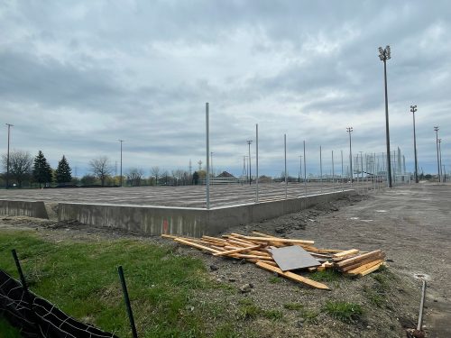 View of the Centennial Park soccer hub under construction, focusing on the excavation work for the new fieldhouse building. The concrete foundation is partially complete and rectangular in shape, with metal poles installed vertically on one side. A pile of wooden pickets and boards lay nearby on the dirt-covered construction site. In the background, there are tall stadium light poles and trees visible under a cloudy sky.