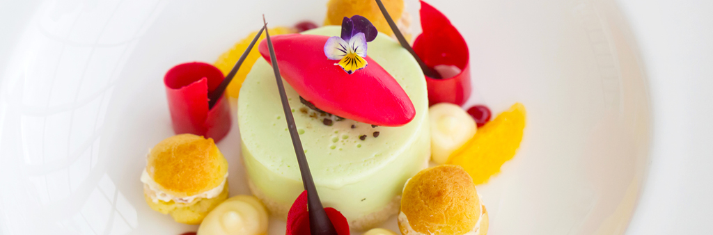 Plated pudding-like desert surrounded by smaller pastries, lines of chocolate and a pansy flower on top