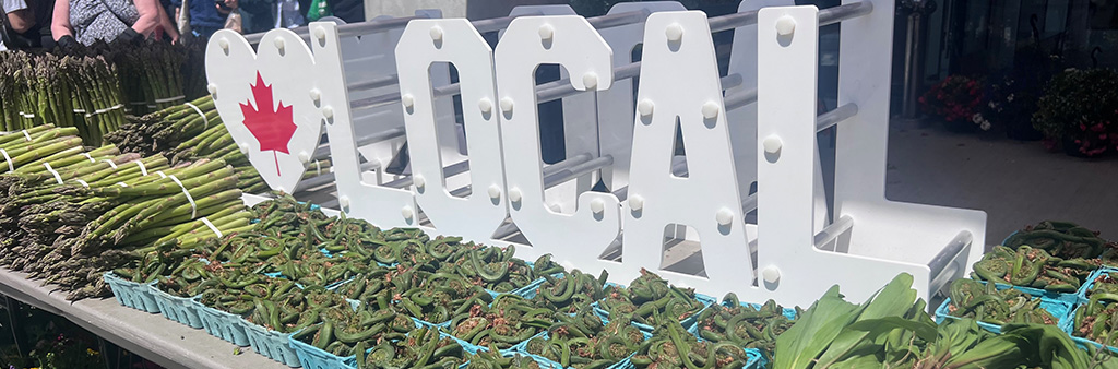 A sign with white block letters that spells LOCAL and starts with a heart sits behind a table filled with fresh produce.