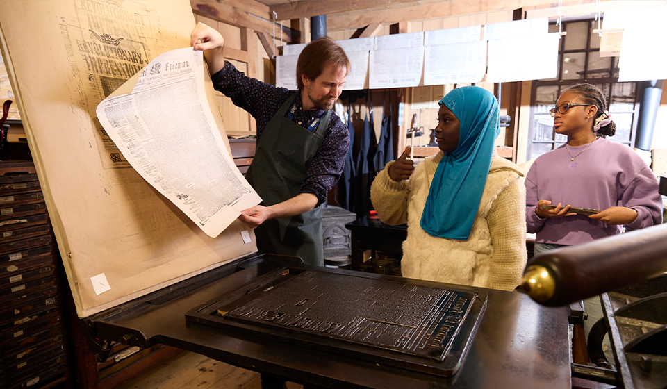 Person holding up a printed sheet stands with two children behind historic printing press