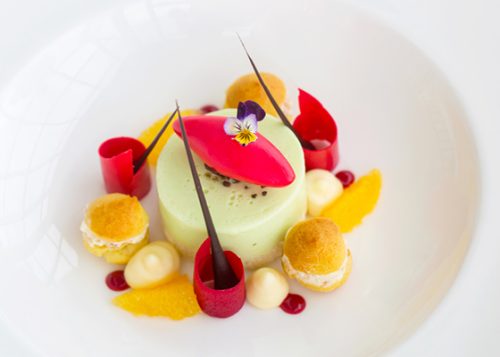 Plated pudding-like desert surrounded by smaller pastries, lines of chocolate and a pansy flower on top