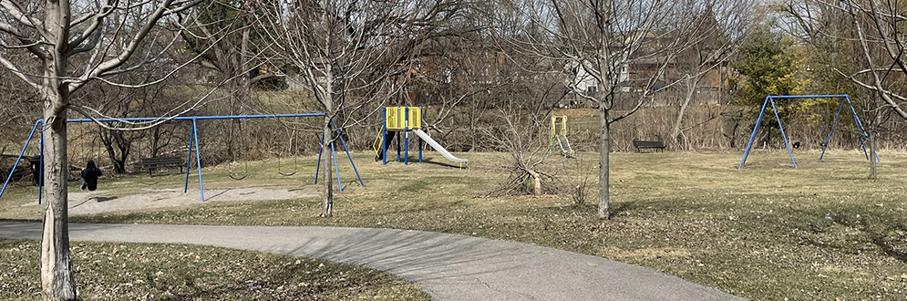 A curved paved path runs through a park with bare trees and a playground in the background. The playground includes blue swing sets and a small slide structure with yellow and green panels. A person is seated on one of the swings. The grass is patchy and brown, with scattered leaves and litter visible on the ground, indicating early spring. Residential houses can be seen in the distance beyond the trees.