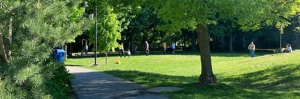 A sunny day at Pricefield Road Playground with people walking and kids playing in an open field surrounded by tall green trees. A paved path curves through the lush grassy area beside pine and deciduous trees.
