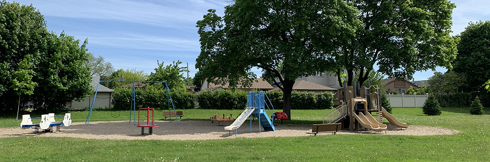 A spacious playground set in a large grassy field, featuring two separate play structures with slides set on sand. The area includes swings, animal-shaped spring riders and a small red climbing structure.