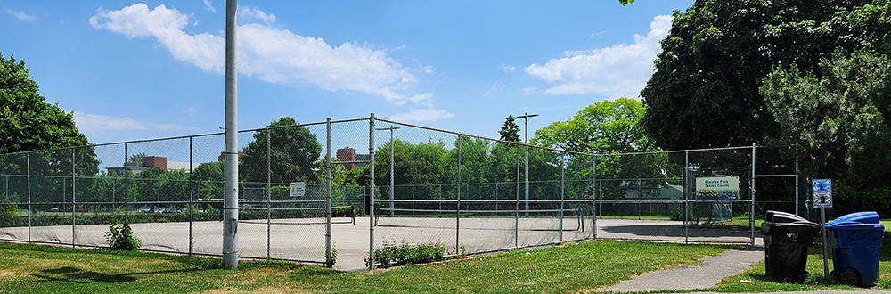 Photo of the existing tennis courts at Clairlea Park on a sunny day, viewing at an angle from the opposite side of a paved accessible pathway. The tennis court is fenced and surrounded by lush green trees. There is lighting to the left and two trash bins to the right of this image.