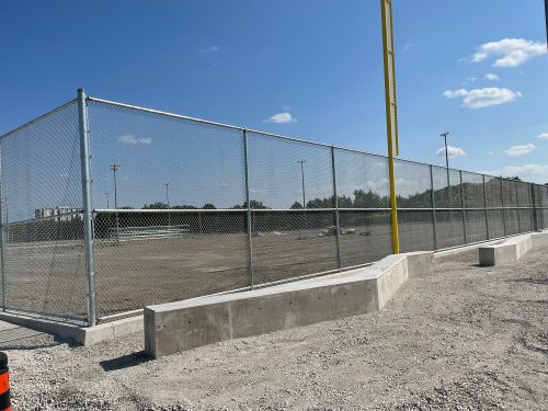 A chain-link fence encloses a large dirt baseball field, with bleachers and light poles.