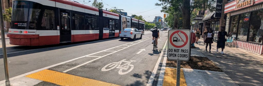 A person cycles in the College Street cycle tracks across an integrated bike/streetcar platform. A streetcar is in the background. A sign at the edge of the platform reads "Do not pass open doors"