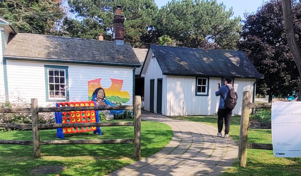 Two white historic houses with mural and connect four game on lawn. Person on path takes photograph with phone.