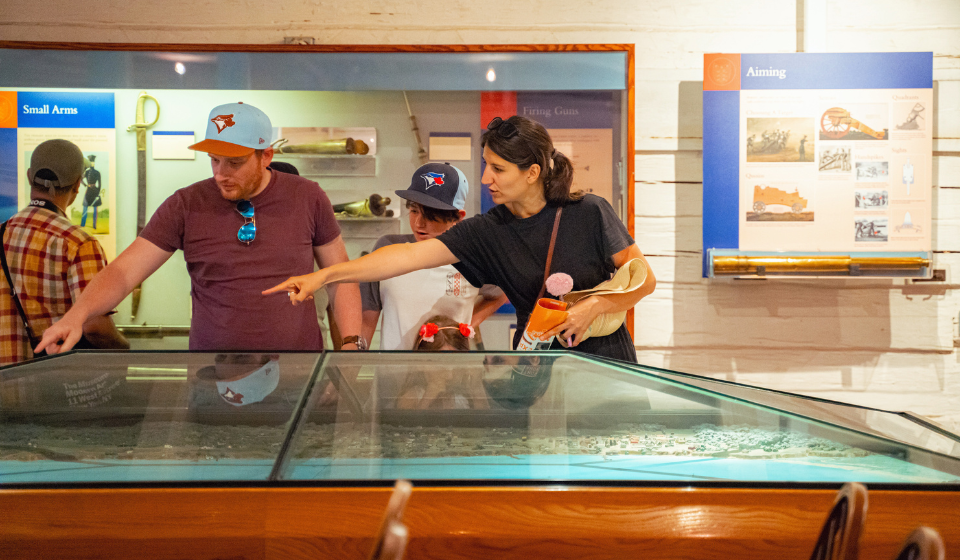 Two adults point to horizontal exhibit case as two children look on. Person in background looks at wall hung exhibits on small weaponry.