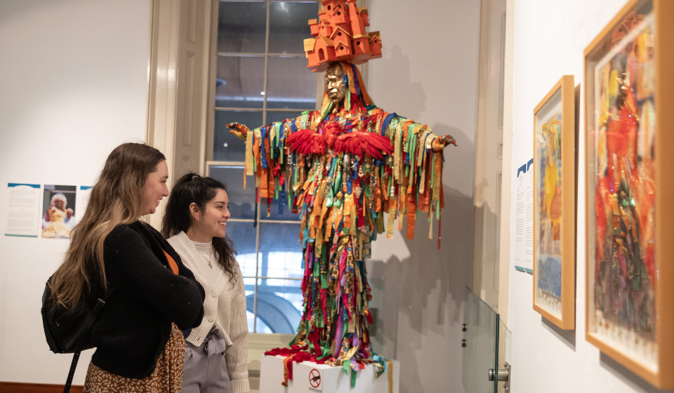Two people in an exhibit gallery look at paintings on wall. Behind them is a sculpture of a person covered in strips of cloth and a miniature building structure on its head.