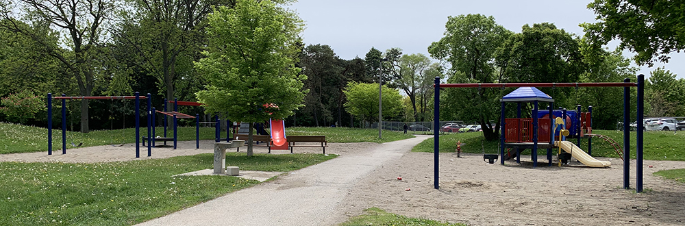 Rowena Park Playground with monkey bars and a small play structure on the right on top of sand, and a larger play structure on the left. The playground is divided by a pathway with mature trees and grass in the background.