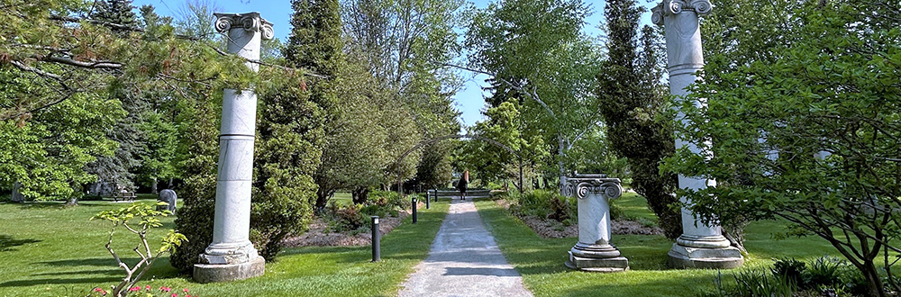 A scenic pathway in Guild Park and Gardens, lined with lush greenery, blooming flowers, and classical marble columns. The path is framed by tall trees and leads toward a metal archway in the distance under a bright blue sky.