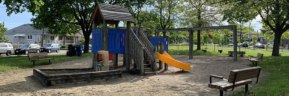 A small neighborhood playground shaded by mature trees, featuring a wooden play structure with a blue panel and orange slide, a sandbox area, and several swings on a wooden frame.