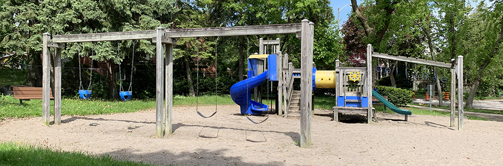 A small playground with wooden structures and a sand surface, featuring a blue spiral slide, green straight slide, climbing elements, and swings. Two baby swings and two belt swings are on the left, and additional swings are on the right.