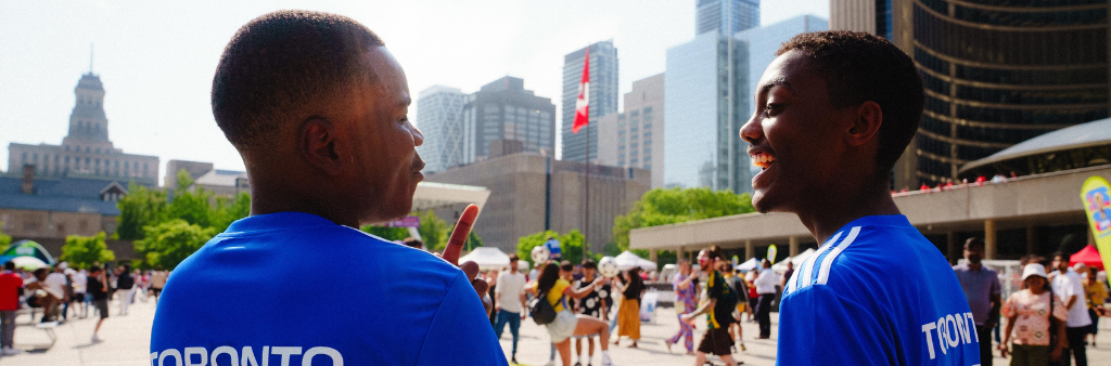 Two people talking and smiling in blue jerseys that read "Toronto" across the back outside Toronto City Hall with a crowd of people in the background.