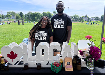 2 people standing behind a farmers' market stall selling local goods in a park.