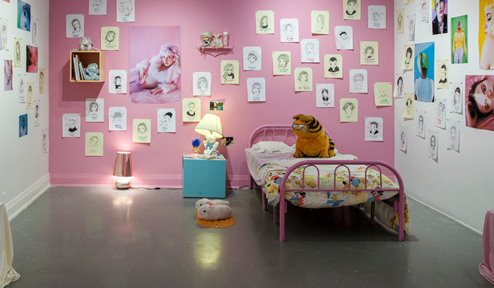 Industrial room decorated as a bedroom with bed, stuffed animals, slippers, desk and lamp and walls covered in artwork