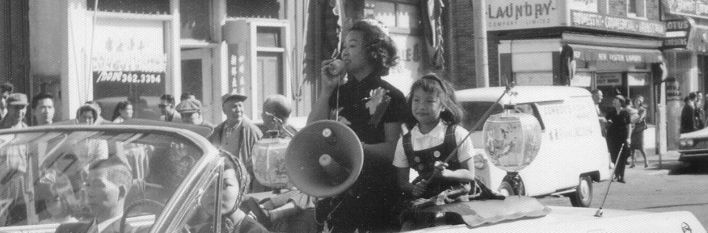 Black and white photograph of woman using a megaphone seated in seated in a convertible car driving through Chinatown