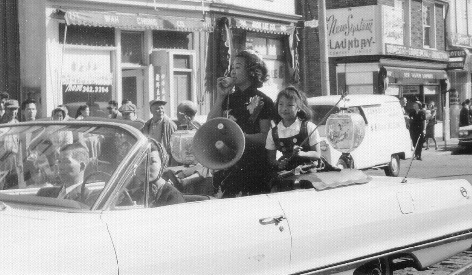Black and white photograph of woman using a megaphone seated in seated in a convertible car driving through Chinatown