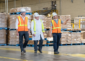three people in high visibility vests walking through a warehouse with palettes loaded with goods.