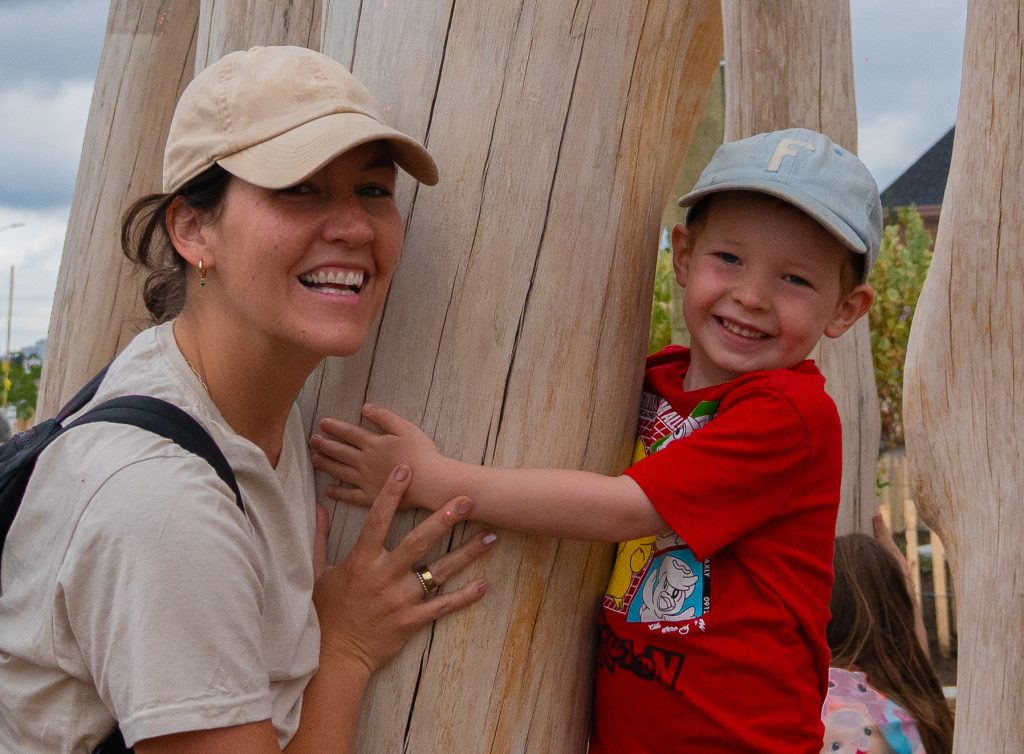A caregiver and child smile while hanging onto the wooden supports of the mock beaver dam.