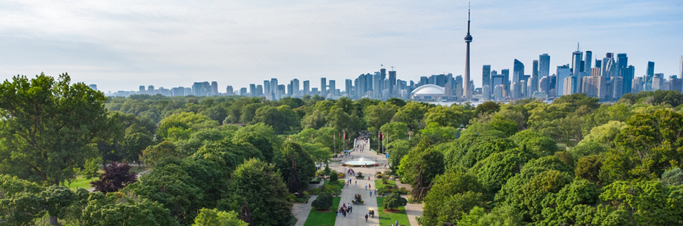 A pathway and a decorative fountain surrounded by trees with the Toronto skyline in the background.
