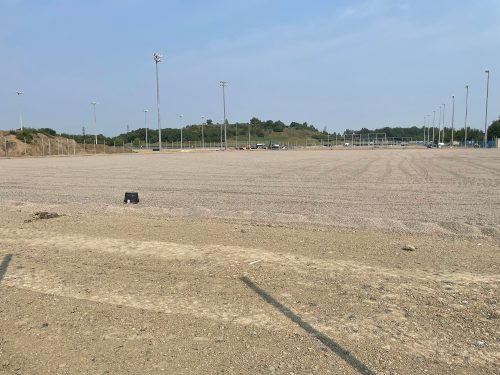 The bubbled training site at the new soccer hub showing the installations in progress. Tall light poles and fencing are installed along the perimeter of the large gravel-covered area.