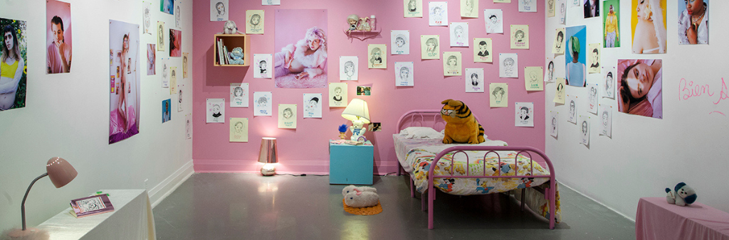 Industrial room decorated as a bedroom with bed, stuffed animals, slippers, desk and lamp, and walls covered in artwork