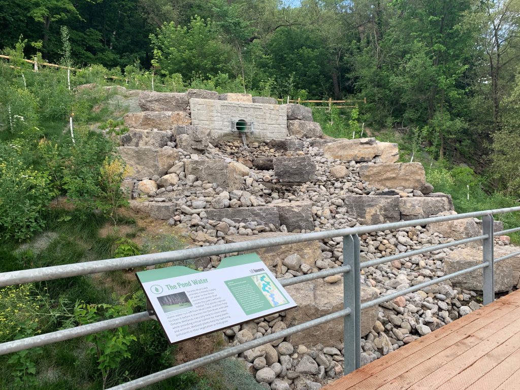 Mud Creek Inlet water cascade feature as originally built in 2020 at Don Valley Brick Works Park. The photo shows a rocky water cascade feature outletting water down a slope, and under a new boardwalk. There are larger and smaller rocks that make up the cascade. New trees and shrubs have been planted around it.