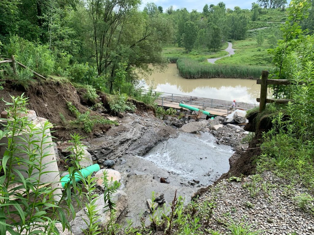 Mud Creek Inlet damage after the July 2024 flooding event. The photo was taken the top of bank off the Belt Line Trail. The photo shows a large break in the embankment, and a jumble of displaced materials from the inlet, including rocks and broken culverts. Damage is significant and the inlet is not in operation.