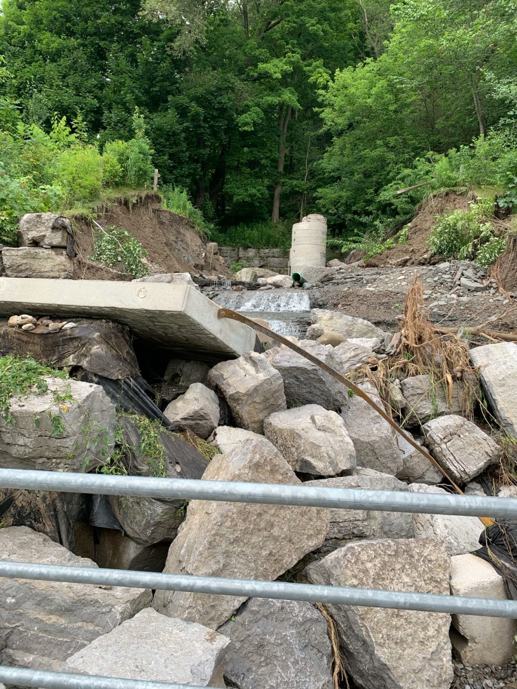 Mud Creek Inlet damage after the July 2024 flooding event, taken from the boardwalk below. The photo shows a large break in the embankment, and a jumble of displaced materials from the inlet, including rocks and culverts. Damage is significant and the inlet is not in operation.