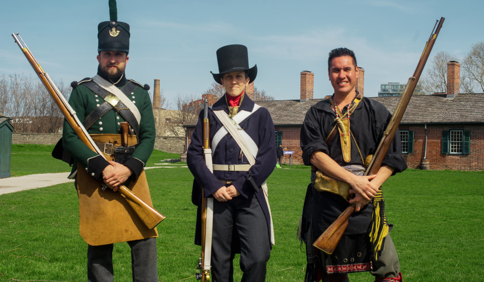 Three historical soldiers in different uniforms stand in field with historic building behind