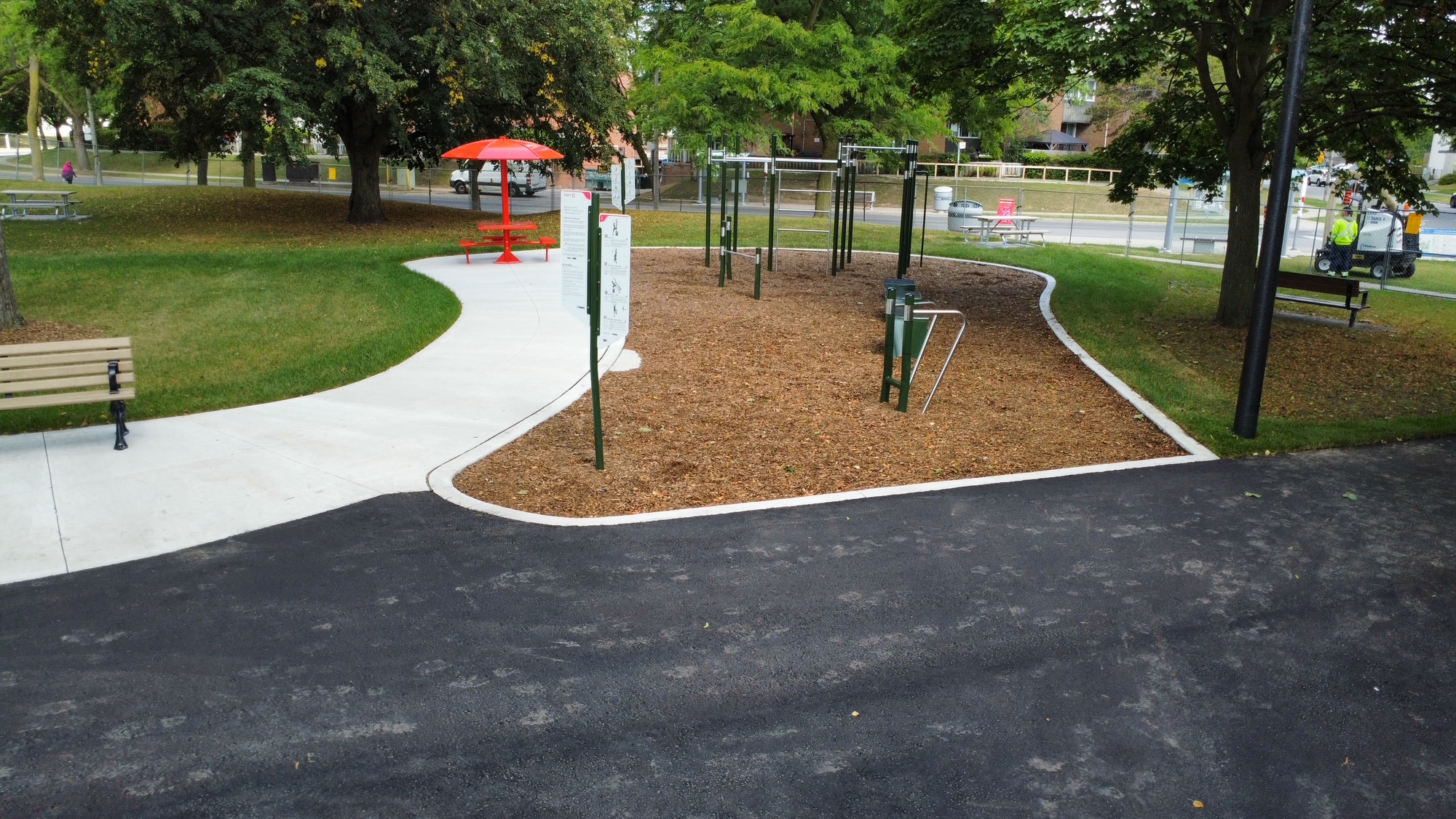 Close-up of outdoor exercise equipment, including bars of varying heights, and accompanying information panels on a mulch surface. Benches and picnic tables are placed on the paved paths and grass lawns surrounding the area.