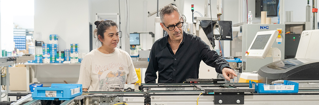 two people standing behind a manufacturing tool inside a plant.