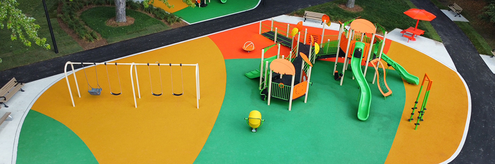 Aerial view of the improved northeast playground area, focusing on the new senior and junior playgrounds, seating areas, and pathways. The playgrounds are brightly coloured and surrounded by trees providing shade.