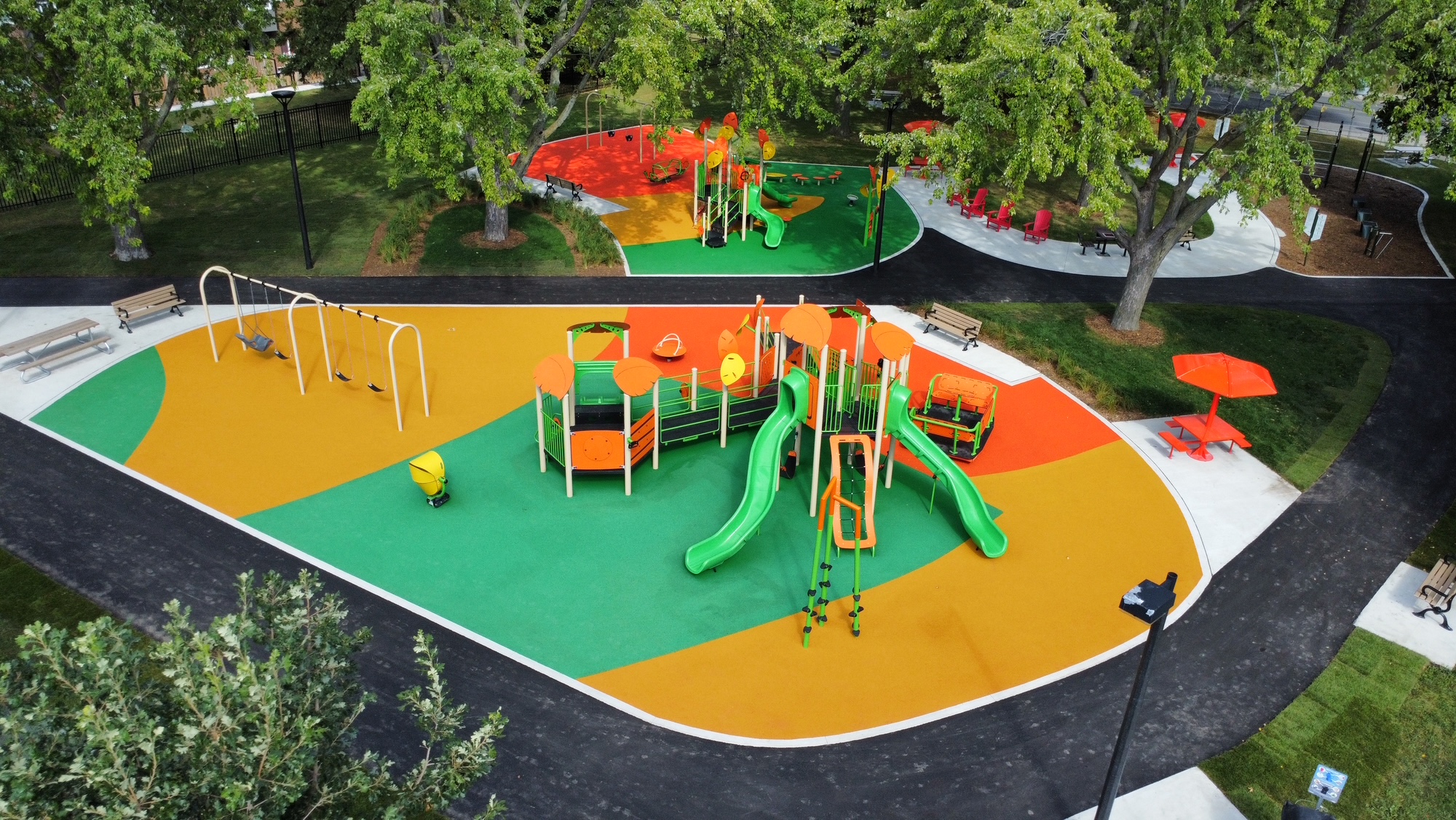 Aerial view of the improved northeast playground area. The playgrounds are separated by a path and include equipment, such as slides and swings, on safety surfacing in a green and orange colour scheme.