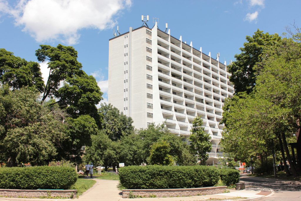 A sunny day in summer featuring a circular parkette space situted within a roundabout with shrubery and mature trees. In the background is a white, rectangular mid-rise apartment building with curved panels projecting from the balconies. 